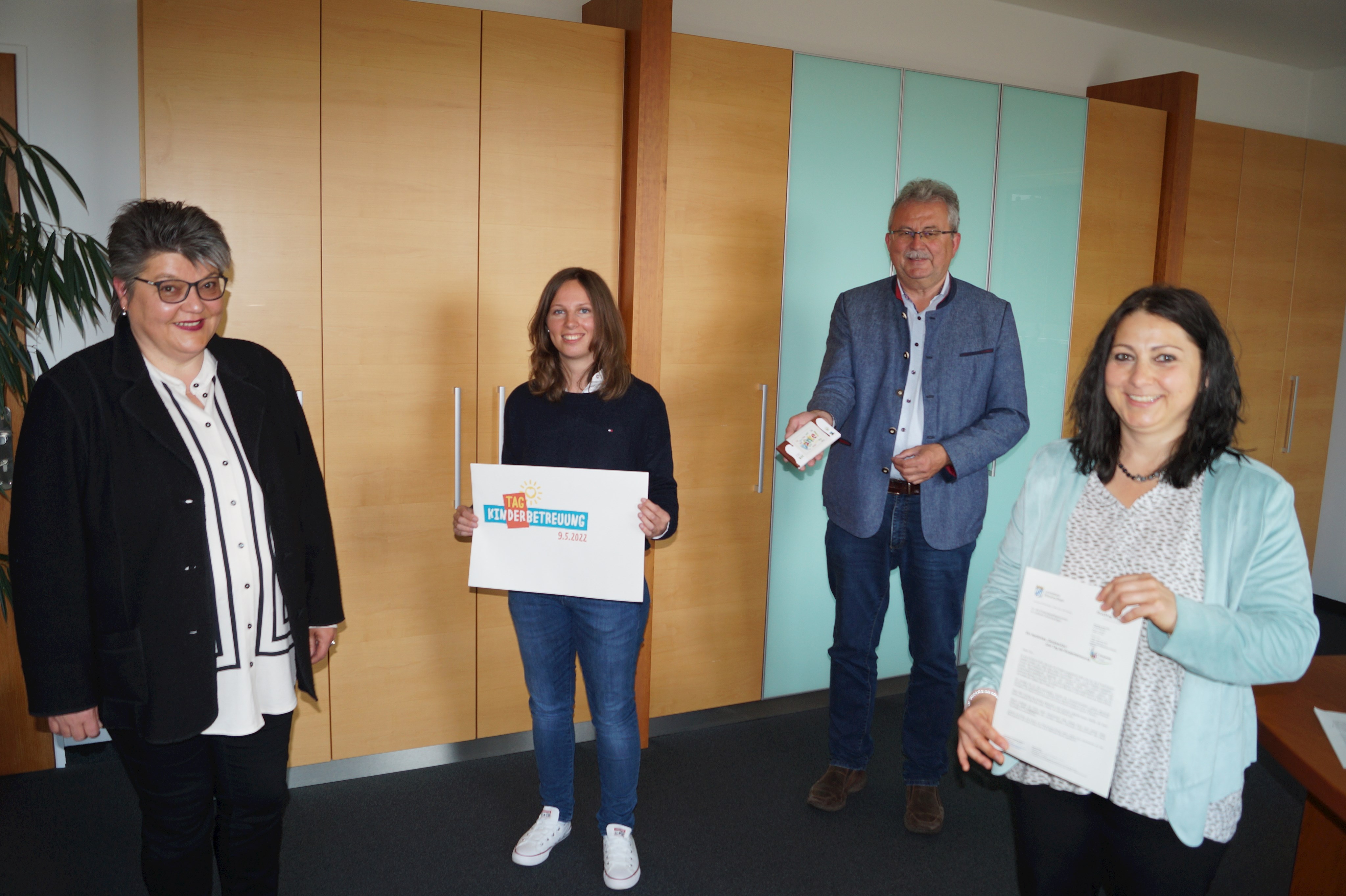 Gerlinde Gietl (Leiterin Amt für Jugend und Familie), Teresa Beck (Amt für Jugend und Familie, Landrat Josef Laumer und Claudia Heißner (Amt für Jugend und Familie, von links) zeigen das Logo zum Tag der Kinderbetreuung im Büro des Landrats.