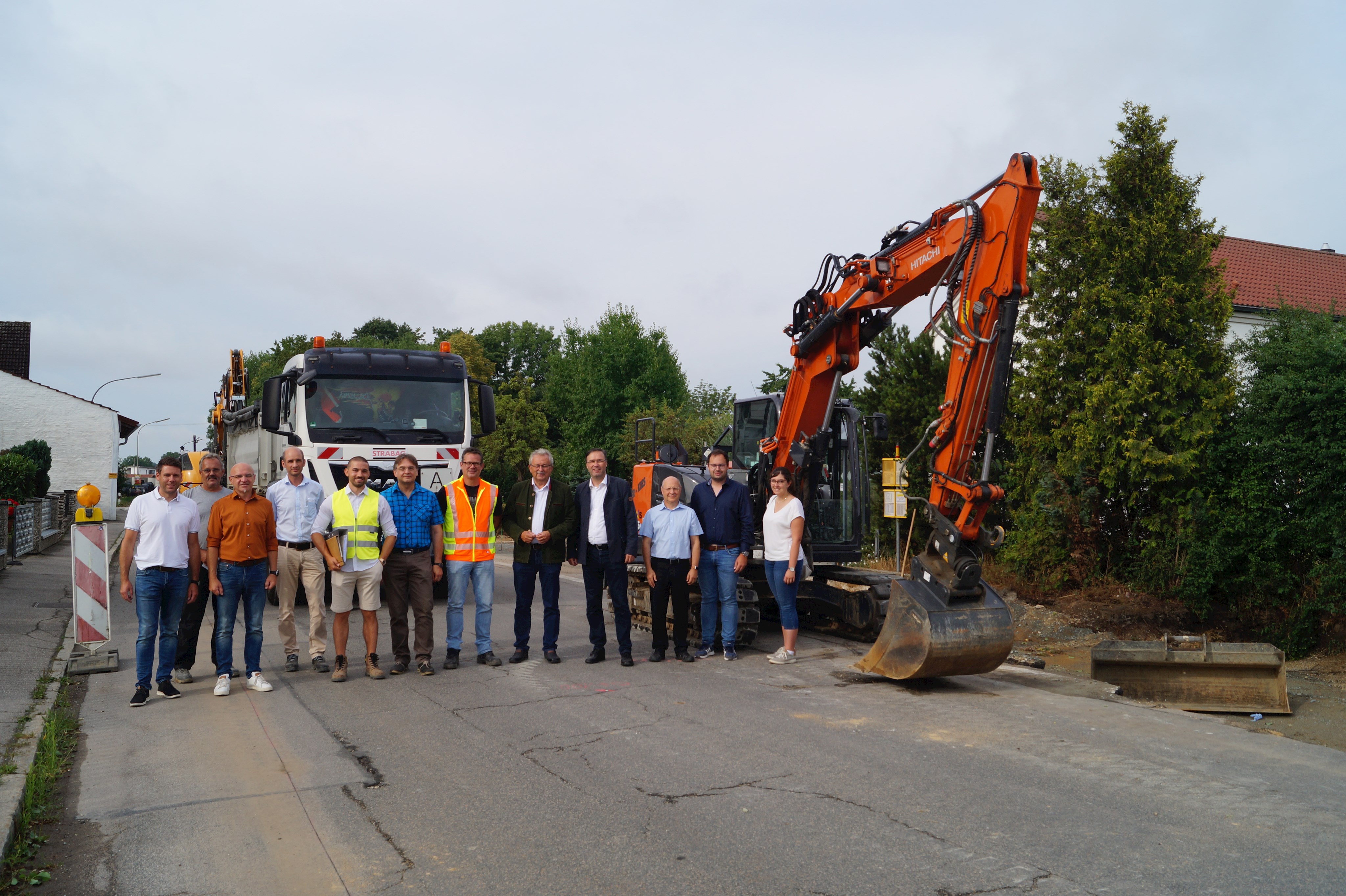 Die Vertreterinnen und Vertreter von Politik, Verwaltung und den beteiligten Firmen eröffneten offiziell die Baustelle an der SR 62 in Parkstetten und stehen auf Höhe der Kirche vor einem Bagger.