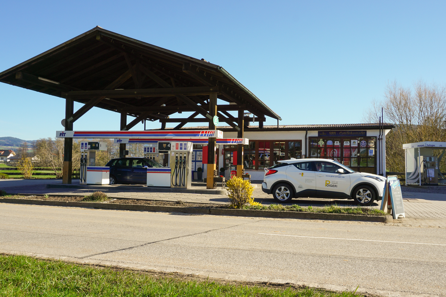 Zu sehen ist die Tankstelle FIT in Hunderdorf von der anderen Strassenseite aus aufgenommen.
Im Hintergrund das Tankstellengebäude, davor die Zapfsäulen. Ein weisses Fahrzeug mit Werbeaufdruck steht am rechten Bildrand. Ein blaues Fahrzeug steht links zwischen den Zapfsäulen.