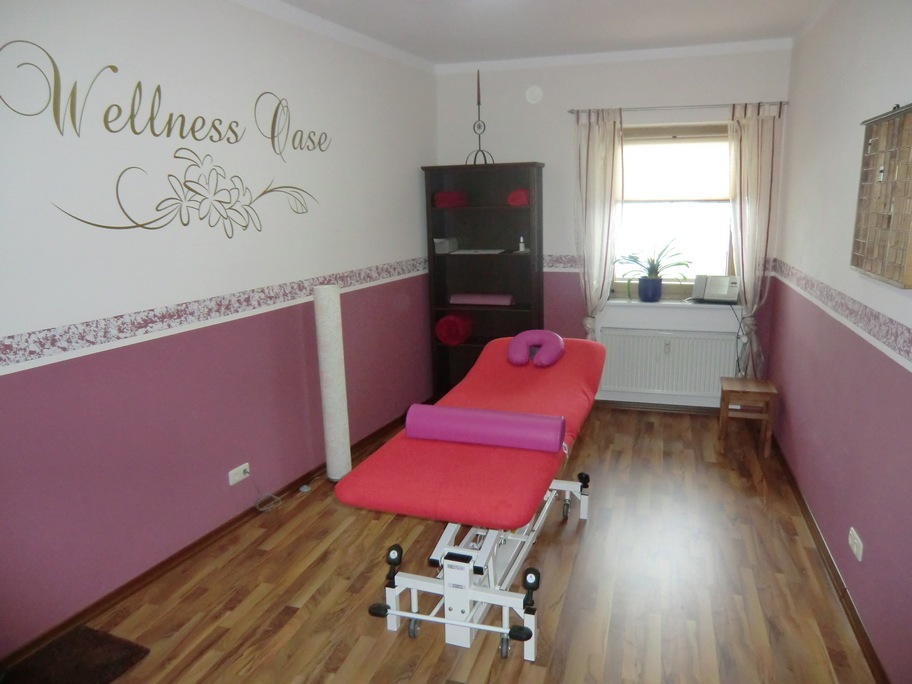 Zu sehen ist ein Behandlungsraum mit zweifarbigen Wänden und dem Schriftzug Wellness Oase an der linken Wand. In der Bildmitte steht eine Massageliege.