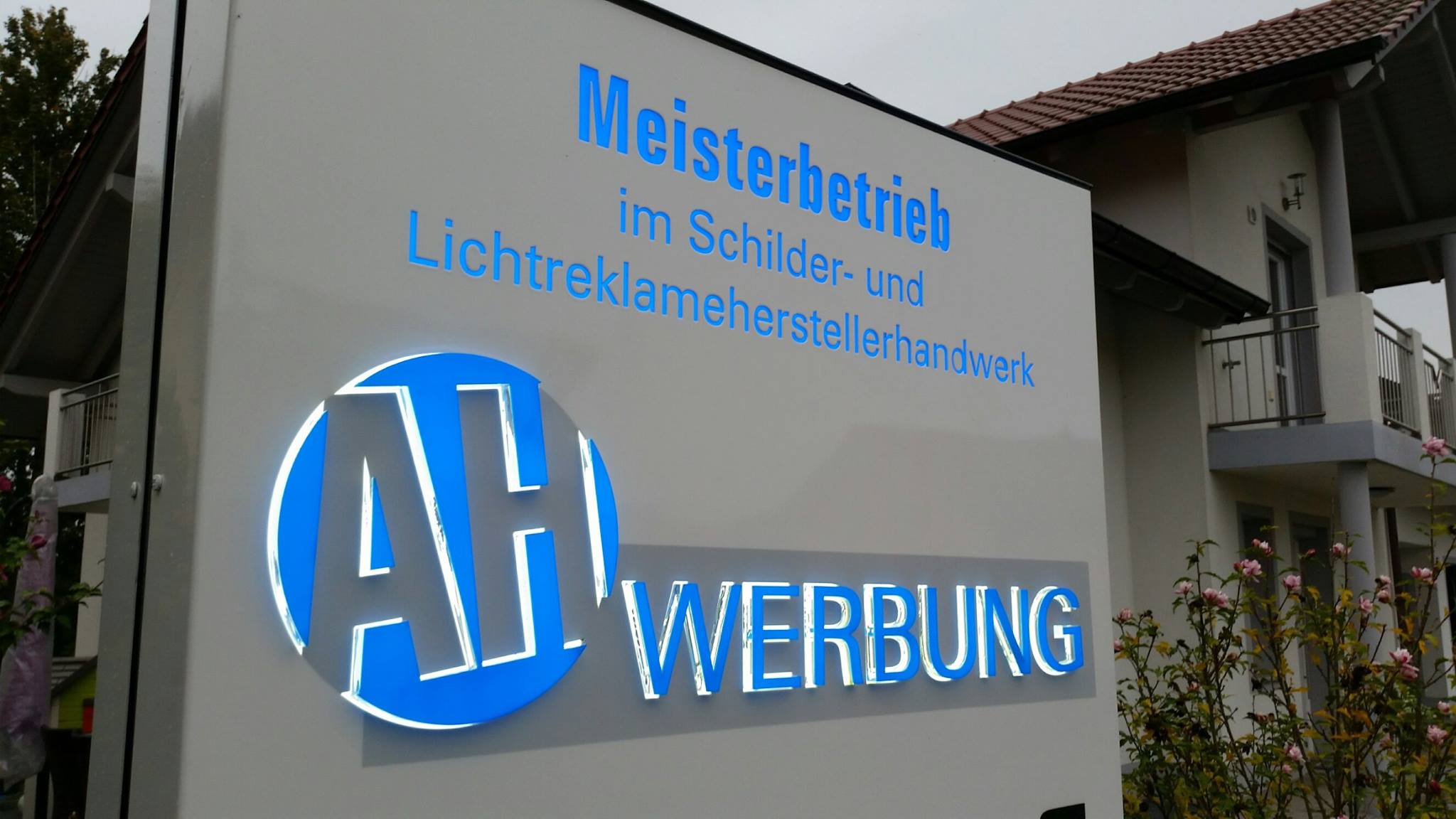 Firmenschild der Firma AH Werbung mit der Aufschrift Meisterbetrieb im Schilder- und Leuchtreklameherstellerhandwerk.
Darunter ist das Logo der Firma zu sehen.