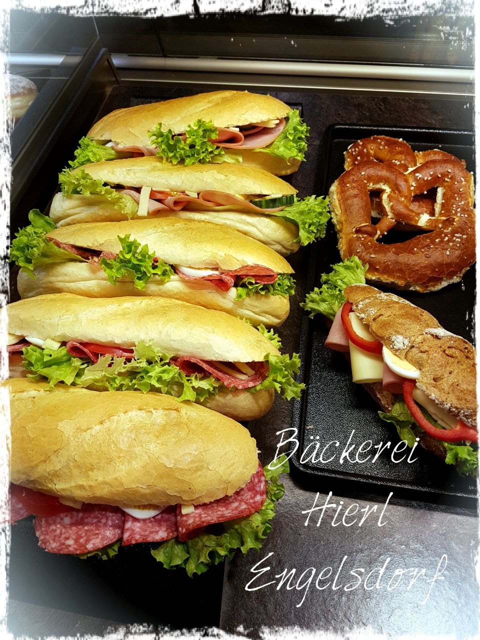 Links sind vier belegte Semmeln abgebildet, rechts daneben ein Brezenstangerln mit Käse und Tomaten, darüber eine Butterbreze.
Rechts unten in der Ecke steht Bäckerei Hierl, Engelsdorf