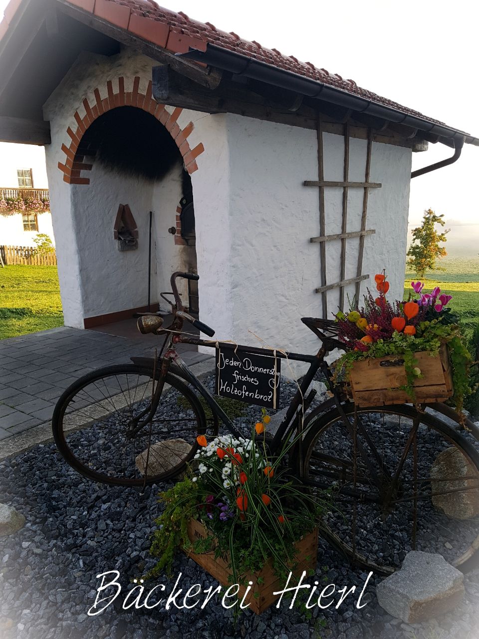 Holzbackofen mit einem alten Fahrrad dafür.
An der oberen Stande des Fahrrades wurde ein Schild mit der Aufschrift Jeden Donnerstag frisches Holzofenbrot befestigt. Auf dem Gepäckträger und vor dem Fahrrad steht eine Kiste mit Blumen.