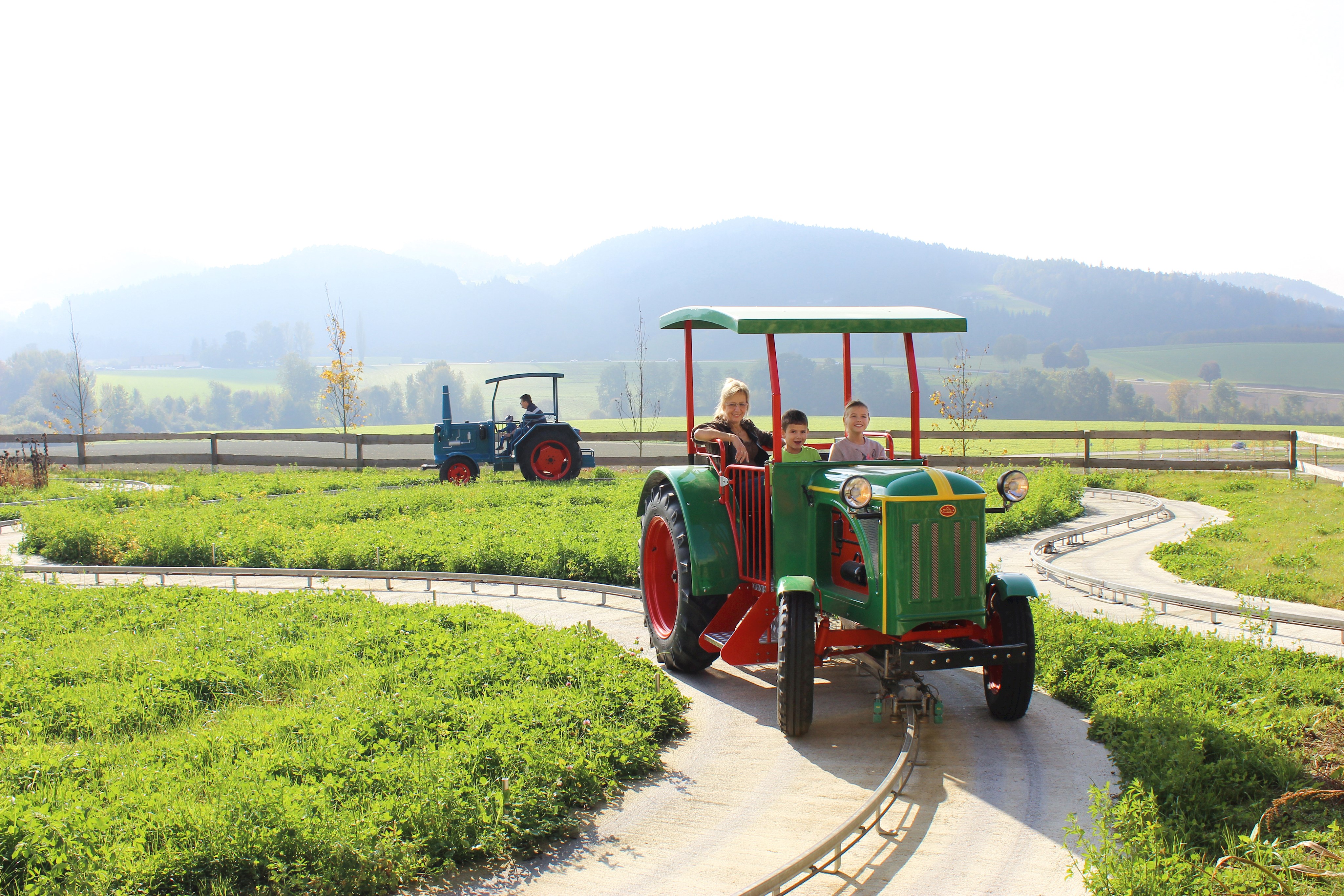 Fahrgeschäft mit Nachbauten alter Traktoren. Im Vordergrund der Nachbau eines alten Deutztraktors in Grün mit zwei Kindern und einer Frau. Im Hintergrund ein blauer Hanomag