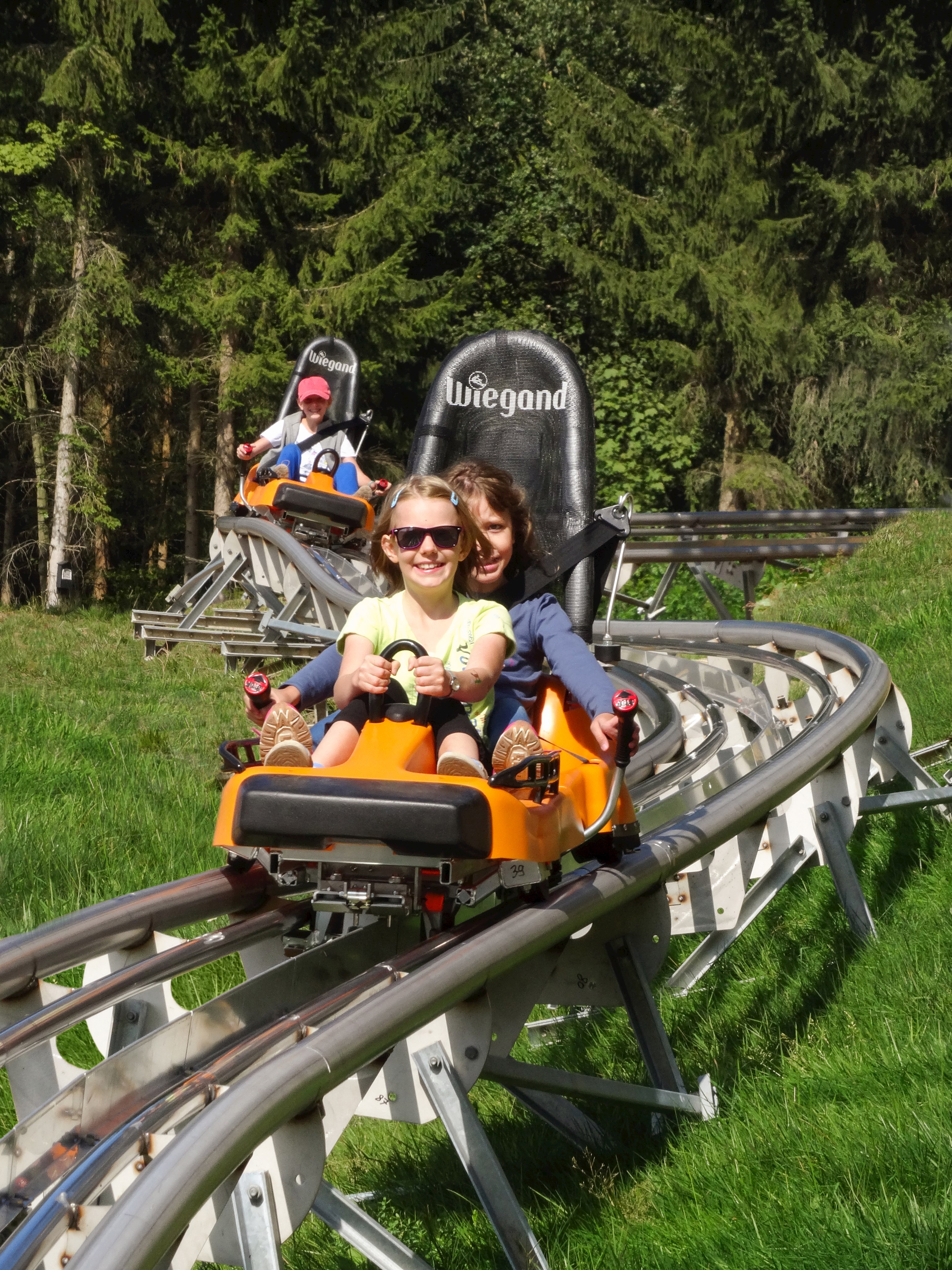 Sommerrodelbahn auf Schienen, im Vordergrund ein oranger Rodel mit zwei Mädchen, dahinter ein weiterer Rodel mit einem Jungen.