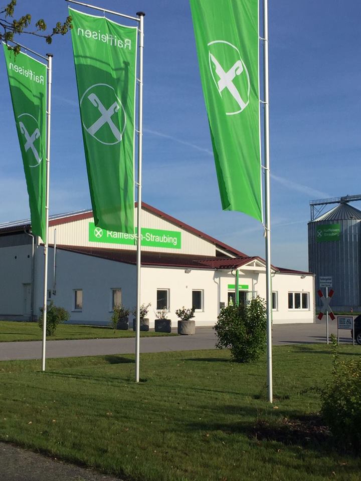 Gebäude der Raiffeisen GmbH im Hafen Straubing. Im Vordergrund drei Masten mit grünen Fahnen auf denen das Raiffeisen-Logo zu sehen ist.