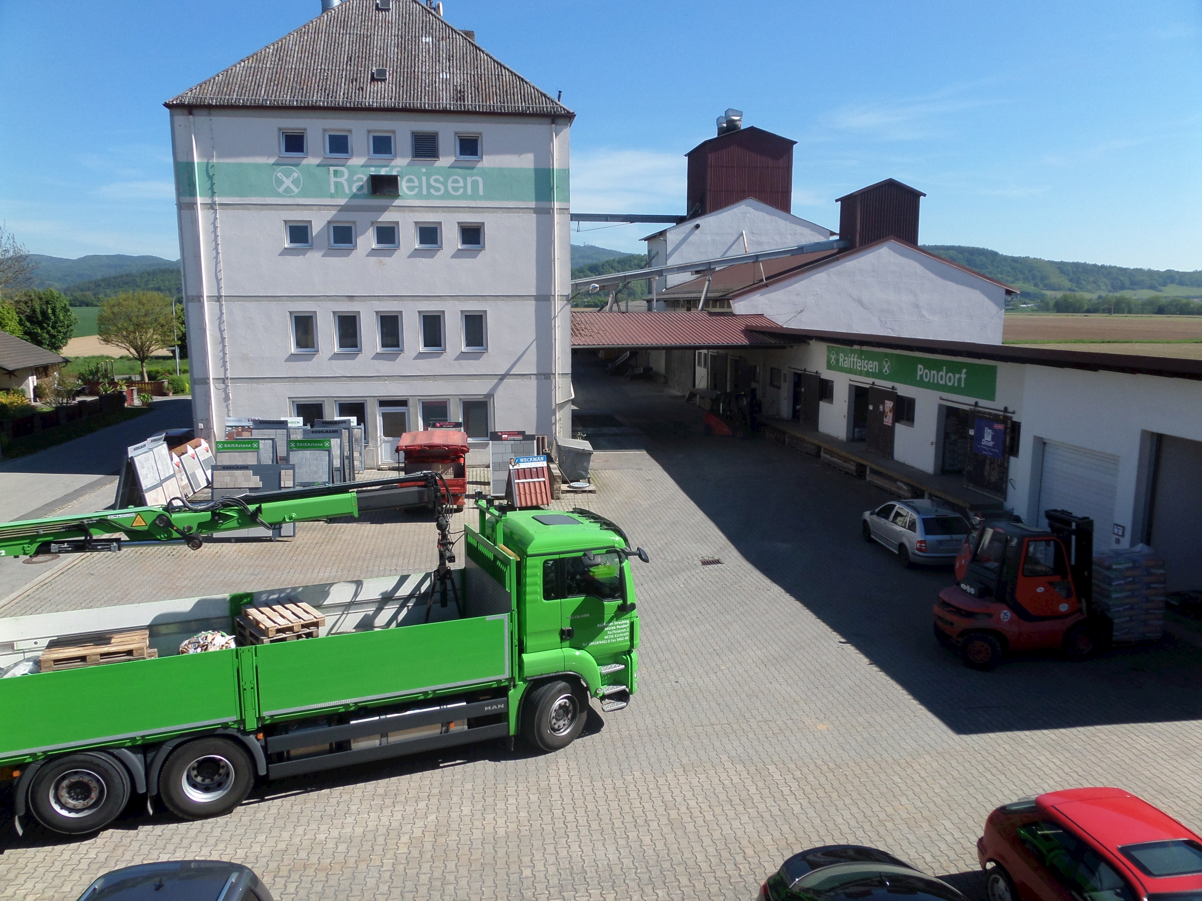 Gebäude der Raiffeisen GmbH in Pondorf. Im Vordergrund ein grüner LKW der Raiffeisen, rechts vor dem Lagergebäude ein Gabelstapler und ein PKW.