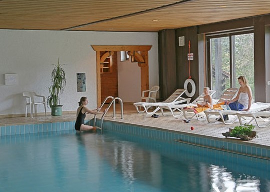 Blick in das Bades des Village Hotels Bayerischer Wald. Rechts mehrere Ruheliegen aus weissem Kunststoff. Ein kleines Mädchen und seine Mutter sitzen jede auf einer Liege. Eine Frau im Badeanzug steigt gerade über die Leiter aus dem Becken.