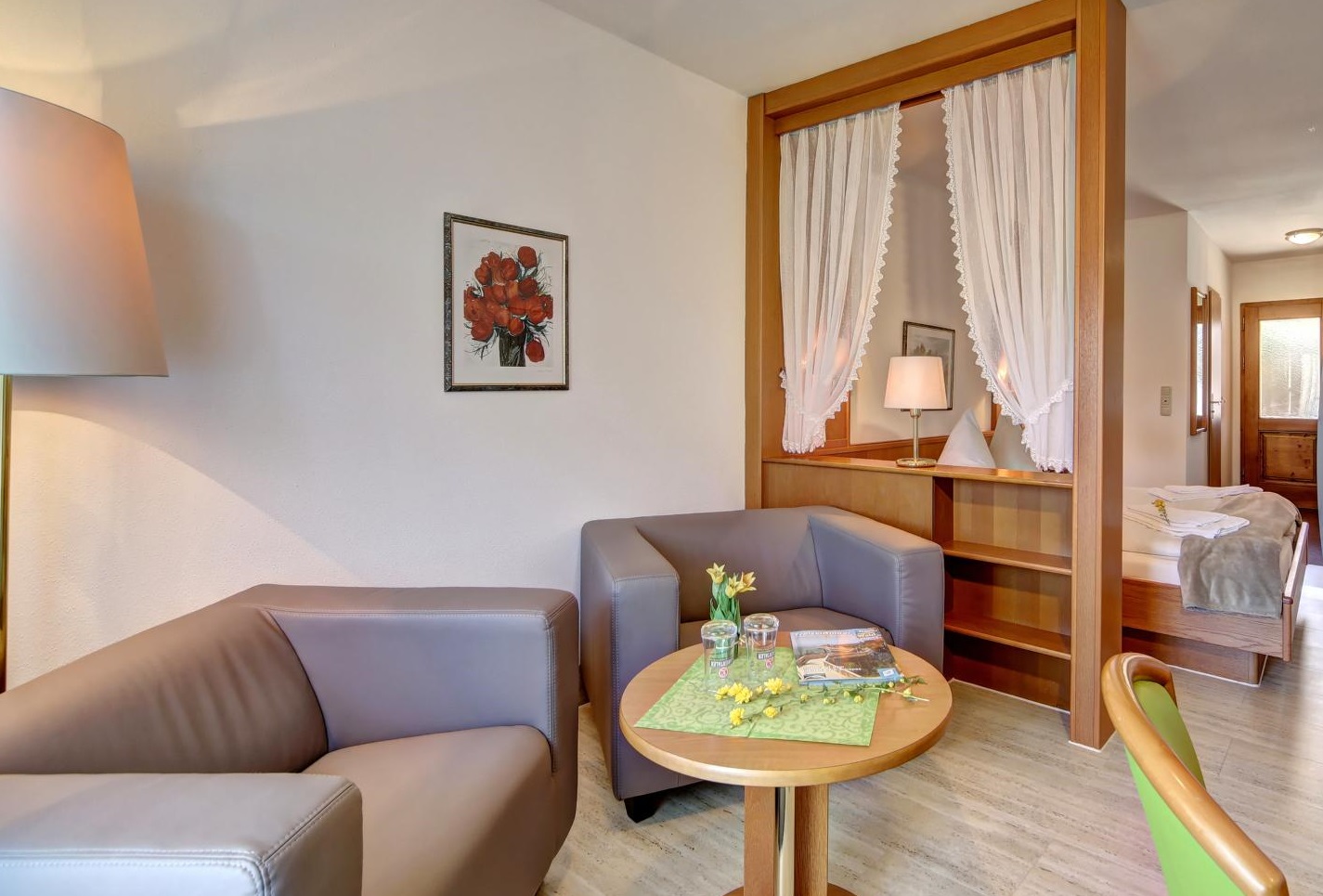 Blick auf die Sitzecke in einem Zimmer. Neben einem runden Holztisch stehen zwei rechteckige Sessel mit Lederbezug. Rechts im Bild sieht man hinter dem Raumteiler das Doppelbett und die Eingangstür. An der Wand hängt ein Bild von einem Blumenstrauß.