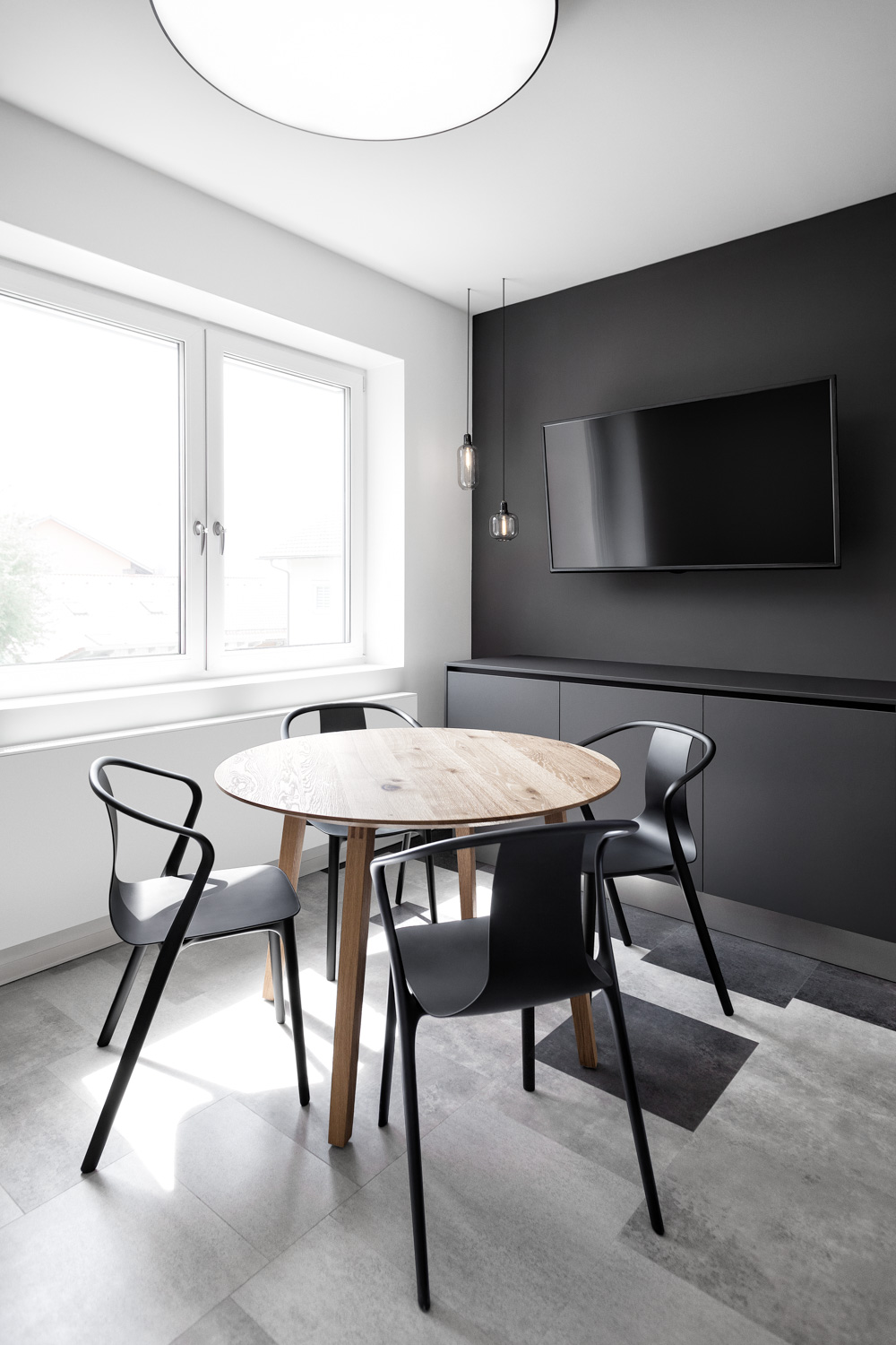 Um einen runden Holztisch stehen vier schwarze Holzstühle. Wand auf der rechten Seite ist antharzit gestrichen, davor steht ein Siteboard in der gleichen Farbe. Ein schwarzer Flachbildschirm hängt an der Wand, davor zwei Hängeleuchten. Links das große Fenster.