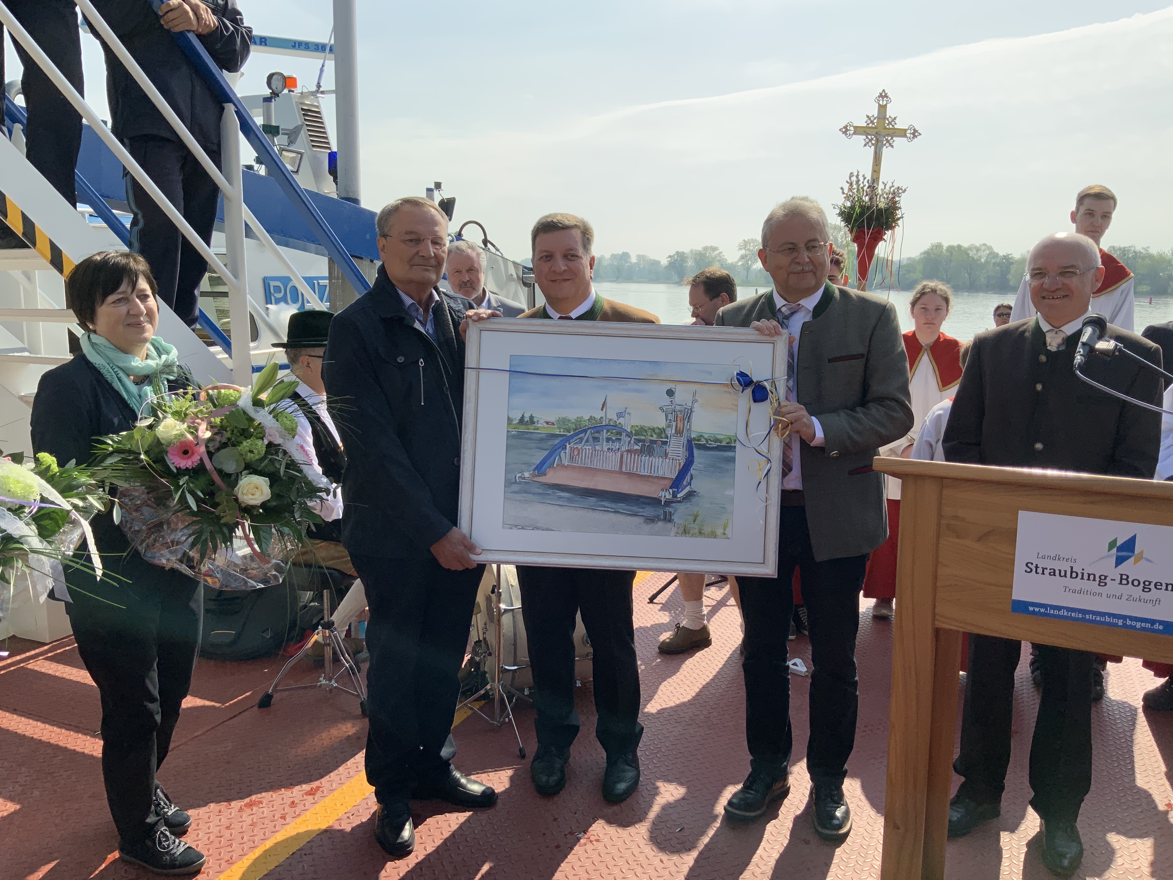 Dr. Dieter Urmann mit seiner Frau, Landrat Christian Bernreiter (Landkreis Deggendorf), Landrat Josef Laumer (Landkreis Straubing-Bogen) und Markus Fischer (Leiter der Tiefbauverwaltung des Landkreises Straubing-Bogen, von links) bei der Geschenkübergabe auf der Fähre Posching mit dem Bild in der Hand.