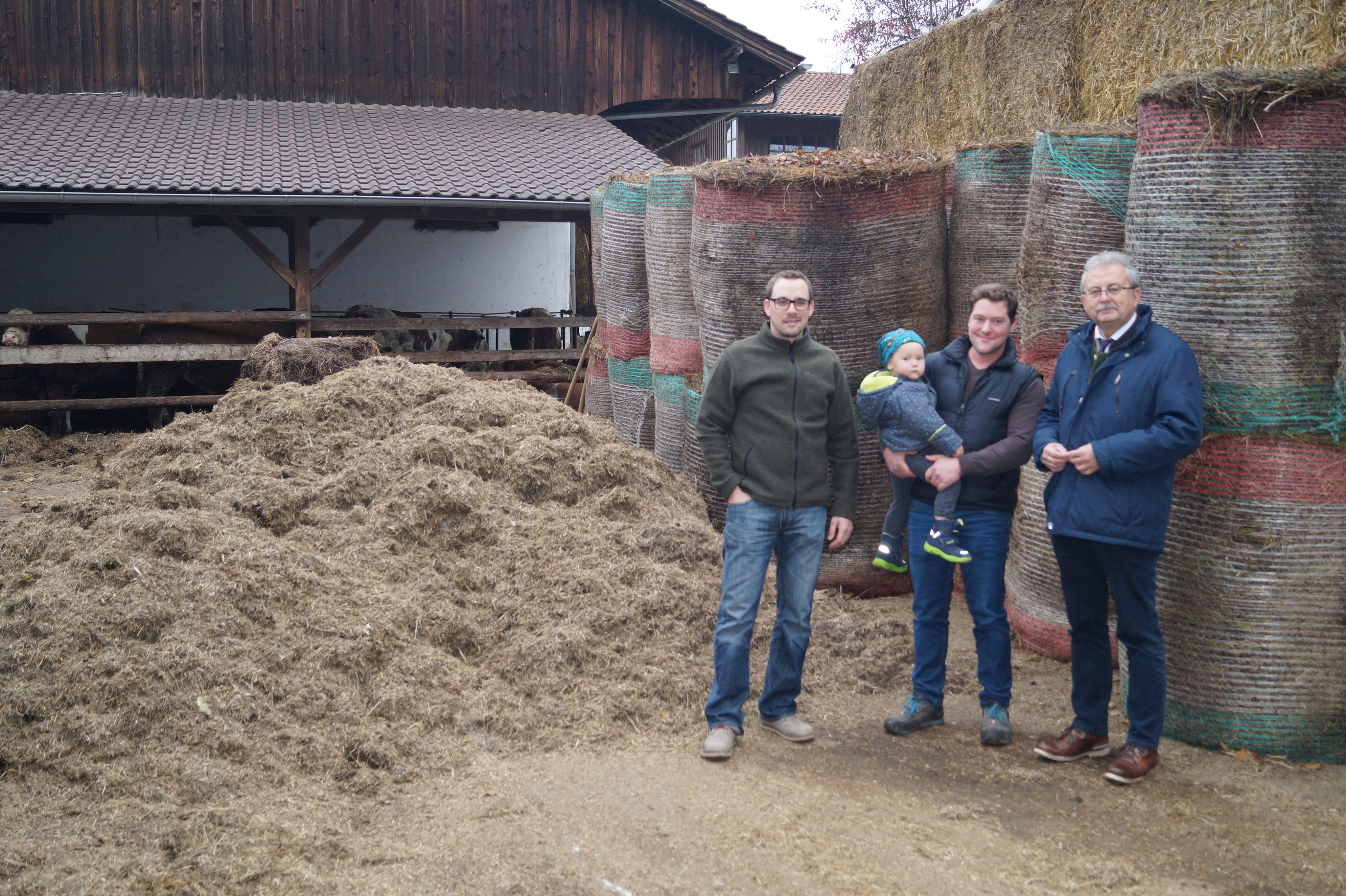 Ambros Köppl vom Landschaftspflegeverband (links) und Landrat Josef Laumer (rechts) ließen sich von Christian Bugl auf seinem Hof aus erster Hand den Rohstoffkreislauf vom Mähgut zum hochwertigen Dünger erklären. Im Hintergrund sind die Einstreu und eine der Stallungen zu sehen.