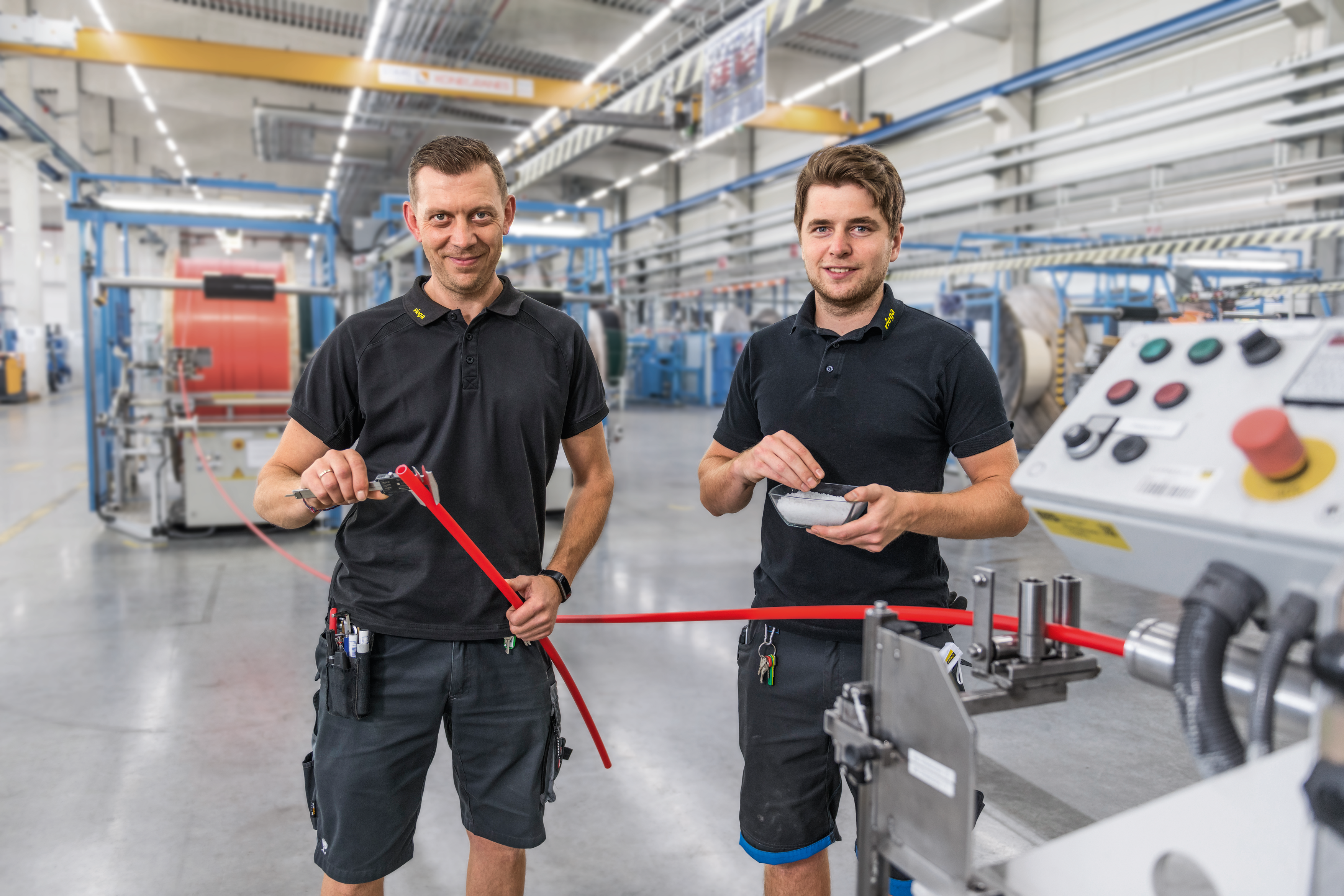 Zwei Mitarbeiter der Firma Viega im Inneren der Produktion. Der Mann links im Bild hält ein rotes Kunststoffrohr in der Hand. Das Rohr läuft aus einer Produktionsmaschine heraus.